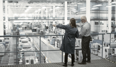 Experienced male and female engineers discuss production at a modern AI-automated electronics manufacturing plant while using laptops.